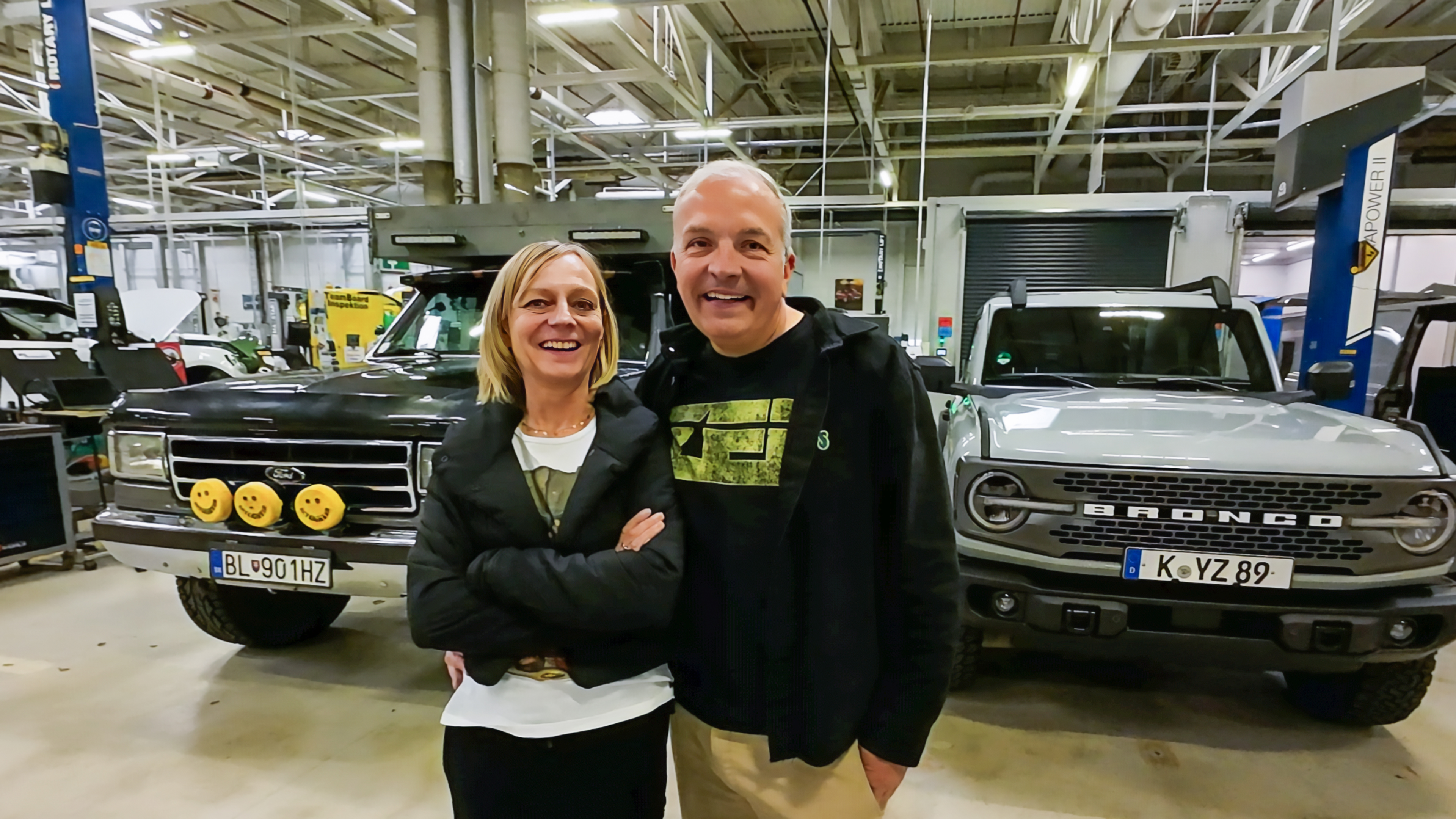 A man and woman pose, smiling, in front of two Ford Broncos in a large garage.